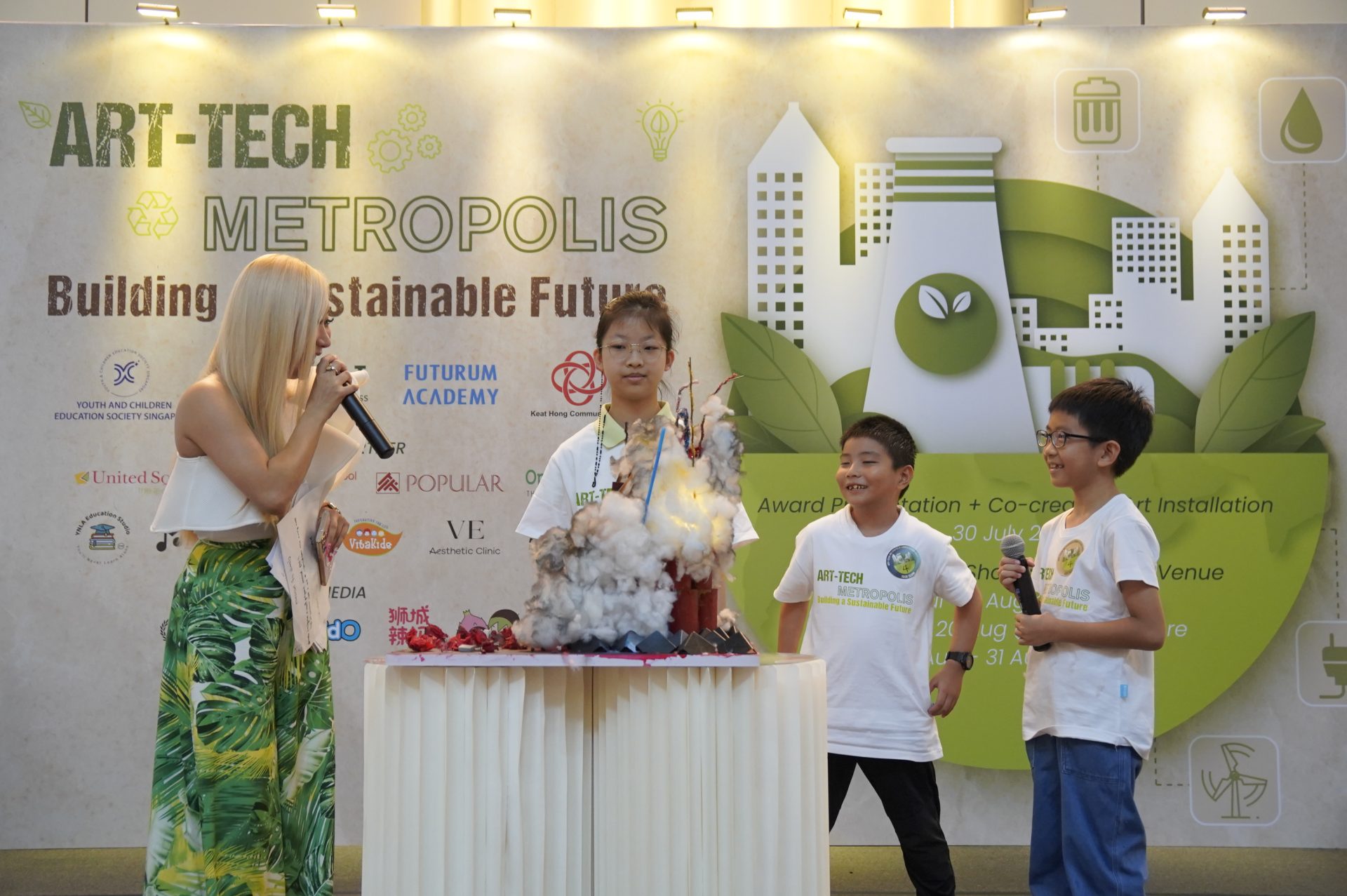 A host with a microphone on stage with three children and their cotton-wool smoke art installation