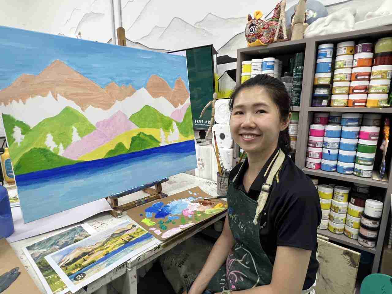 Adult student in a TREE ART apron smiling beside her mountain-lake landscape painting on an easel